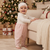 A baby standing indoors next to a cream sofa, wearing a white knit bonnet and a matching white lace-trim top with pale pink footed pants. A decorated Christmas tree and wrapped gifts are visible in the background.
