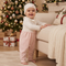 A baby standing indoors next to a cream sofa, wearing a white knit bonnet and a matching white lace-trim top with pale pink footed pants. A decorated Christmas tree and wrapped gifts are visible in the background.