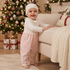 A baby standing indoors next to a cream sofa, wearing a white knit bonnet and a matching white lace-trim top with pale pink footed pants. A decorated Christmas tree and wrapped gifts are visible in the background.