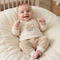 A smiling baby wearing a cream and beige two-piece outfit with a train graphic on the chest, lying on a quilted cream baby lounger.