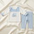 A baby two-piece knit outfit set laid flat on a cream satin fabric background. The set includes a long-sleeve top with a white body, light blue yoke and cuffs, and a blue train graphic on the chest, paired with matching light blue jogger pants with white cuffs.