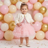 A toddler girl standing in front of a festive pink, cream, and gold balloon background with confetti on the floor, wearing an off-white faux fur cropped bolero jacket over a pink dress with a sequined bodice and tiered ruffled tulle skirt.