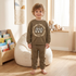 A young child stands barefoot in a bright room wearing an olive brown sweatshirt and matching sweatpants set. The sweatshirt features a circular graphic with the words 'Urban Legends' and 'Dreamers'.