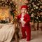 A baby wearing a festive red Christmas pajama set with white lace trim and a matching red hat, standing next to a small wooden stool in a decorated holiday room with a Christmas tree and warm lights.