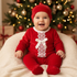 A baby wearing a festive red Christmas outfit with a matching beanie, lace trim, and white bow, sitting on a cream knit chair in front of a decorated Christmas tree.