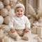 Baby sitting in a neutral-themed room wearing a white and beige knit sweater set and matching beanie.