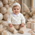 Baby sitting in a neutral-themed room wearing a white and beige knit sweater set and matching beanie.