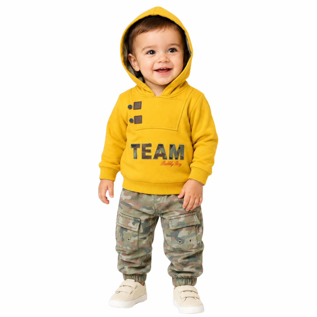 A toddler standing against a plain white background, wearing a bright yellow hooded sweatshirt with 'TEAM' printed across the front and camouflage cargo pants with white sneakers.