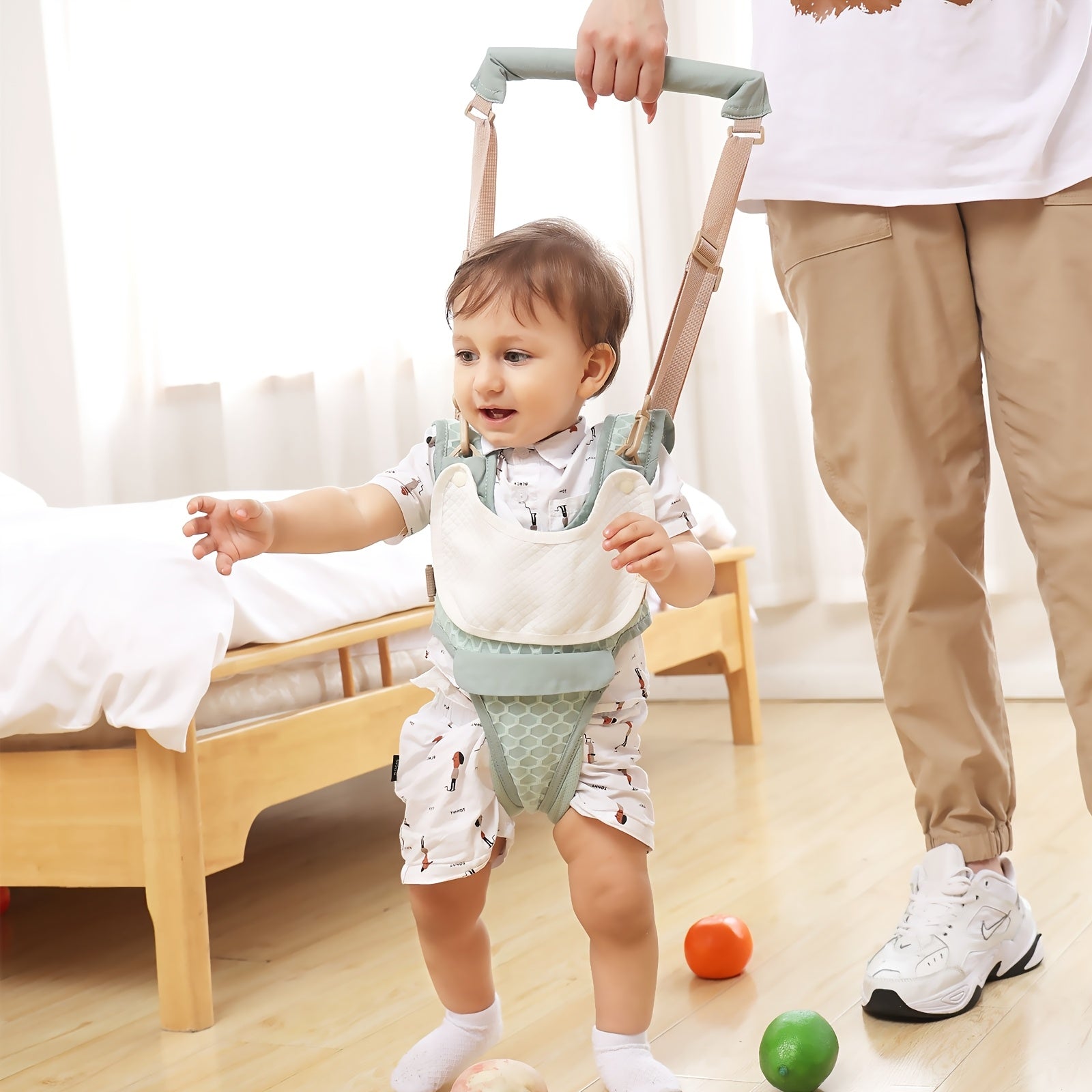 Baby using a walking frame with a person assisting, on a wooden floor with colorful balls.