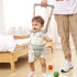 Baby using a walking frame with a person assisting, on a wooden floor with colorful balls.
