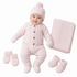 A baby wearing a light pink cable-knit one-piece outfit with a matching knit hat, mittens, booties, and a folded blanket on a plain white background.