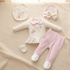 A baby clothing set laid flat on a textured cream bedspread, including a long-sleeve footed romper in off-white with pink collar and bow details, pink leggings with white toe sections, a matching off-white bib, a pink bonnet with a bow, and a small off-white bootie.
