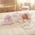 A sleeping newborn baby wearing a pink and white embroidered outfit, lying on a cream quilted mattress with a matching bib and hat nearby. A woven bassinet and soft nursery room are visible in the background.