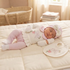 A sleeping newborn baby wearing a pink and white embroidered outfit, lying on a cream quilted mattress with a matching bib and hat nearby. A woven bassinet and soft nursery room are visible in the background.