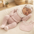 A sleeping newborn baby wearing a dusty pink footed sleeper, matching headband with lace, and a floral embroidered bib, lying on a textured cream blanket.