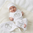 A smiling baby lies on their back on a cream-colored blanket, wearing a white matching set of a cap, bib, short-sleeve bodysuit, and footed pants, all featuring a gray elephant pattern and the text 'welcome to the world'[cite: 1].