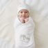A smiling baby wrapped in a white swaddle blanket and matching cap, featuring a gray elephant print and the text 'welcome to the world', lies on a cream blanket[cite: 1].