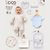 A baby outfit collection display featuring a smiling baby wearing a cream footed sleeper with a blue and yellow pattern, surrounded by matching baby clothing and accessories on a light gray background. The collection includes a light blue short-sleeve bodysuit, a matching bonnet, bibs, a storage pouch, and a branded gift bag.