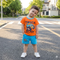 Child wearing an orange shirt with a truck graphic and blue shorts standing on a street.
