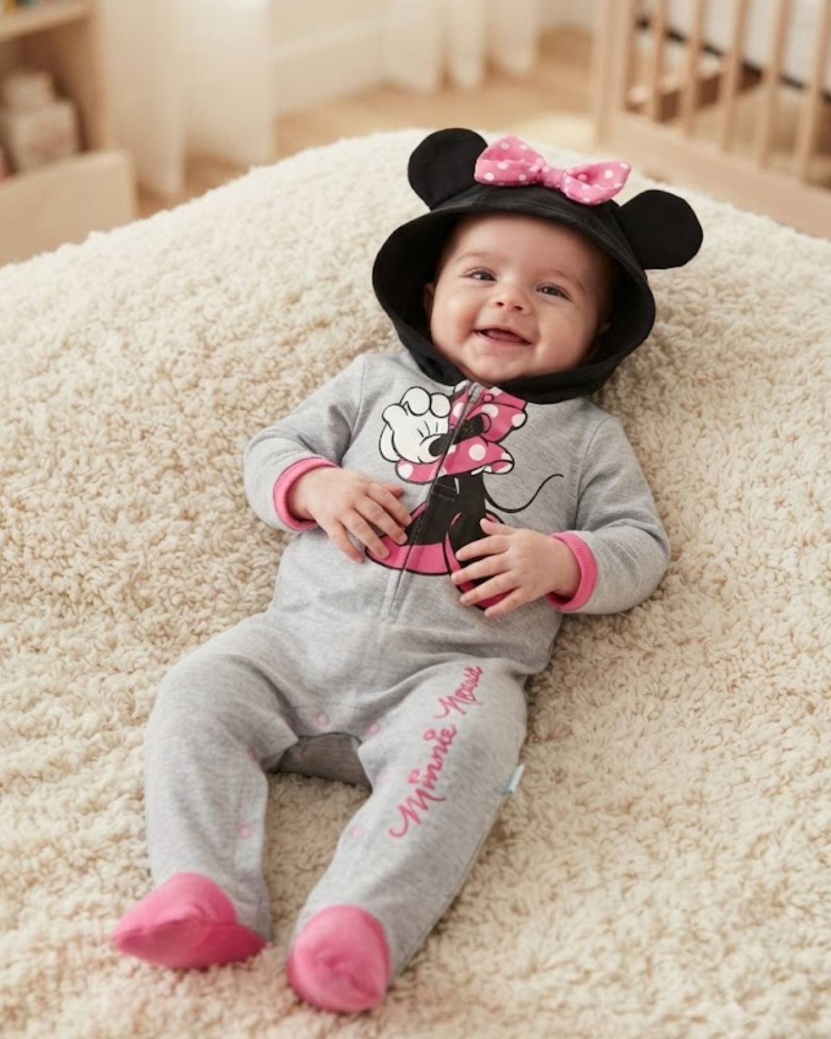 A baby lying on a cream carpet wearing a gray Minnie Mouse onesie with a black and pink Minnie Mouse hat.