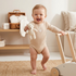Laughing baby in a beige long-sleeve bodysuit holding a mini teddy bear.