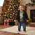 A toddler stands barefoot on a cream knit rug in a cozy holiday living room, wearing a black zip-up puffer jacket over a green graphic t-shirt and blue jeans. A decorated Christmas tree, wrapped gifts, and a fireplace are visible in the background.