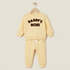 A baby boy's two-piece sweatshirt and jogger pants set in pale yellow, hanging on a wooden hanger against a light beige background. The sweatshirt features the words 'DADDY'S MINI' printed in dark brown across the chest, and the pants have a white drawstring.