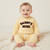 A baby sitting on a cream knit blanket wearing a pale yellow sweatshirt and matching pants with 'DADDY'S MINI' printed in dark brown across the chest.