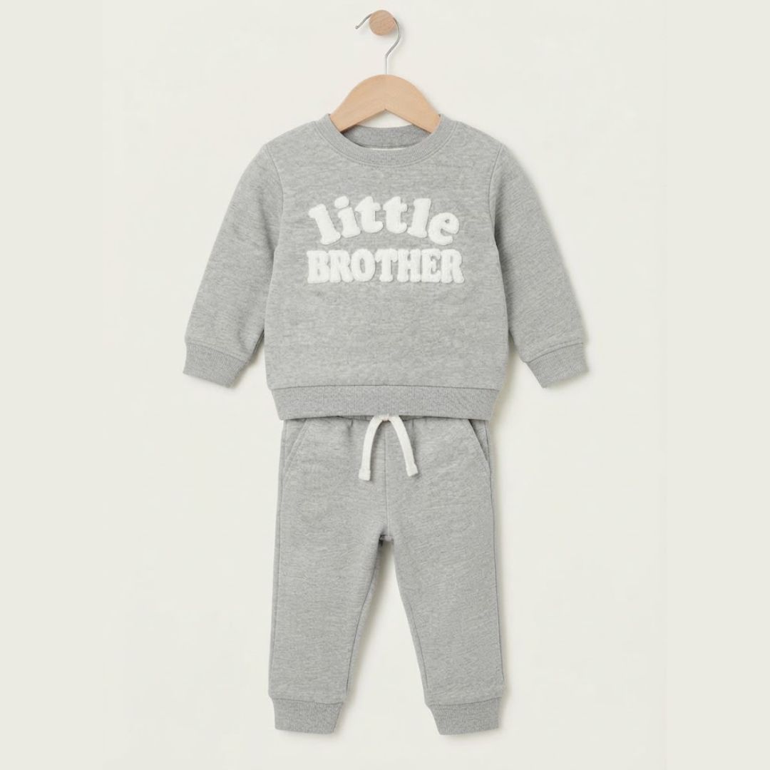 A toddler's two-piece heather gray sweatshirt and jogger pants set hanging on a wooden hanger against a light beige background. The sweatshirt features the words 'little BROTHER' printed in white across the chest.