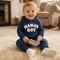 A baby boy sitting on a cream shag rug wearing a navy blue hoodie and matching sweatpants with 'MAMA'S BOY' printed in white across the chest. He is holding a small book in a cozy living room setting.