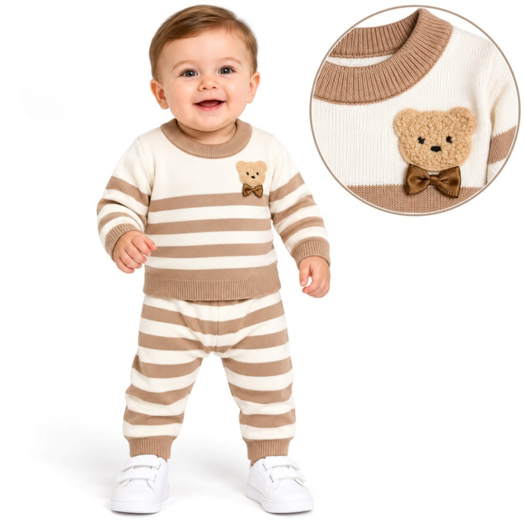 A smiling baby in a brown-and-white striped knit sweater and pants with a fuzzy teddy bear applique, wearing white sneakers