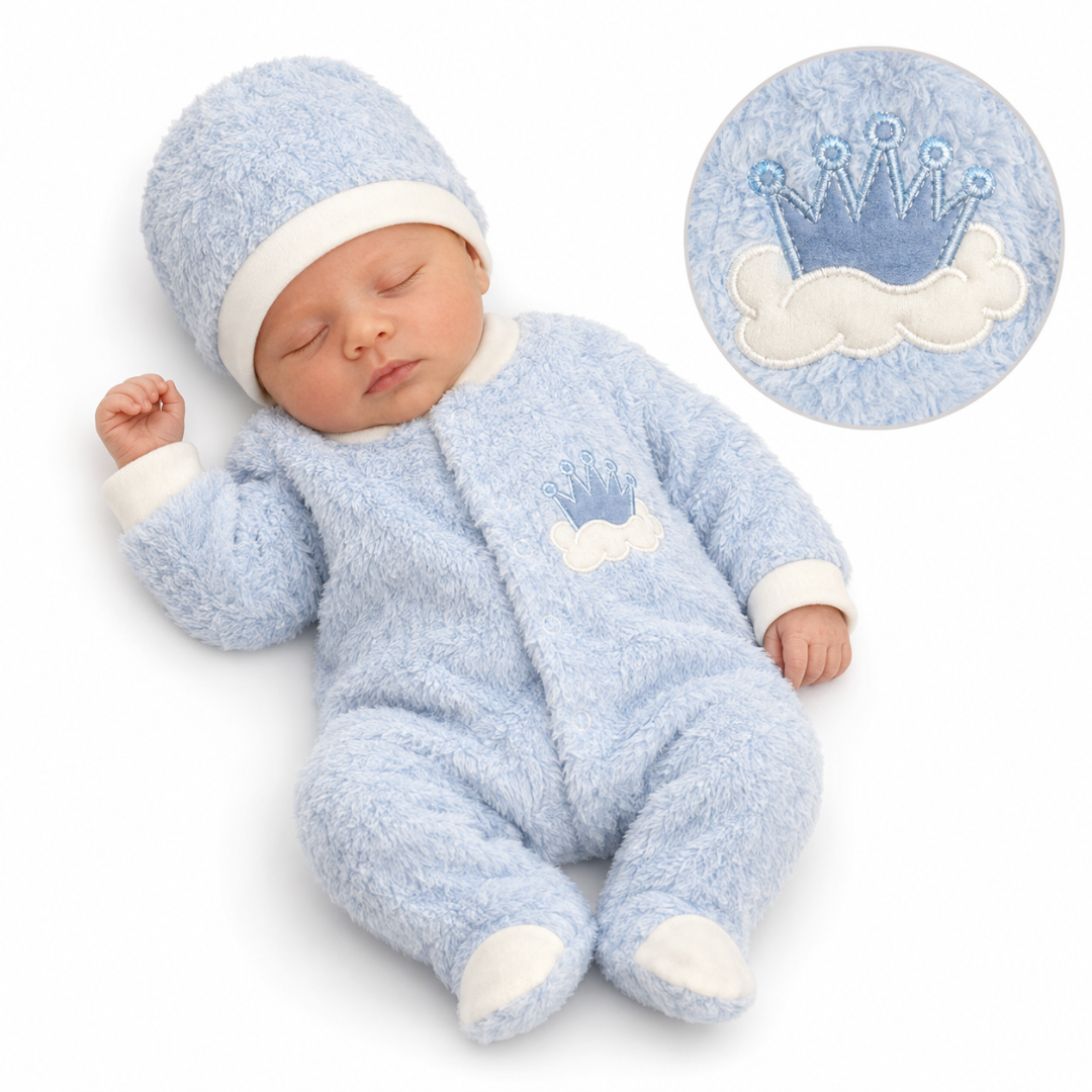 A sleeping newborn baby wearing a light blue fuzzy footed onesie and matching hat with white trim and a crown-and-cloud applique. A circular inset at the top right shows a close-up of the blue-and-white crown motif.