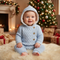 A baby wearing a light blue cable-knit hooded romper sits on a white faux fur rug in front of a decorated Christmas tree and fireplace. The romper has a soft cream lining inside the hood and three gold buttons on the chest.