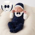 A sleeping newborn baby wearing a navy blue velvet footed romper styled like a tuxedo with a white Peter Pan collar, matching navy bow tie, and a navy-and-white beanie. A circular inset at the top left shows a close-up of the tuxedo-style collar detail.