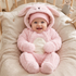 A baby wearing a plush pink bunny costume with a hood and matching hat, sitting in a crib with beige bedding and a stuffed toy in the background.