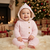 A baby wearing a light pink cable-knit hooded romper sits on a white faux fur rug in a cozy Christmas setting with a decorated tree, fireplace, and wrapped gifts in the background.