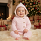 A baby wearing a light pink cable-knit hooded romper sits on a white faux fur rug in a cozy Christmas setting with a decorated tree, fireplace, and wrapped gifts in the background.