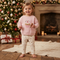 A young child stands barefoot in a cozy holiday room, wearing a light pink sweatshirt with a cupcake graphic and the words 'Santa's Little Helper' and matching cream pants with small pink motifs. A decorated Christmas tree, fireplace, wrapped gifts, and a knitted rug are visible in the background.