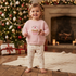 A young child stands barefoot in a cozy holiday room, wearing a light pink sweatshirt with a cupcake graphic and the words 'Santa's Little Helper' and matching cream pants with small pink motifs. A decorated Christmas tree, fireplace, wrapped gifts, and a knitted rug are visible in the background.
