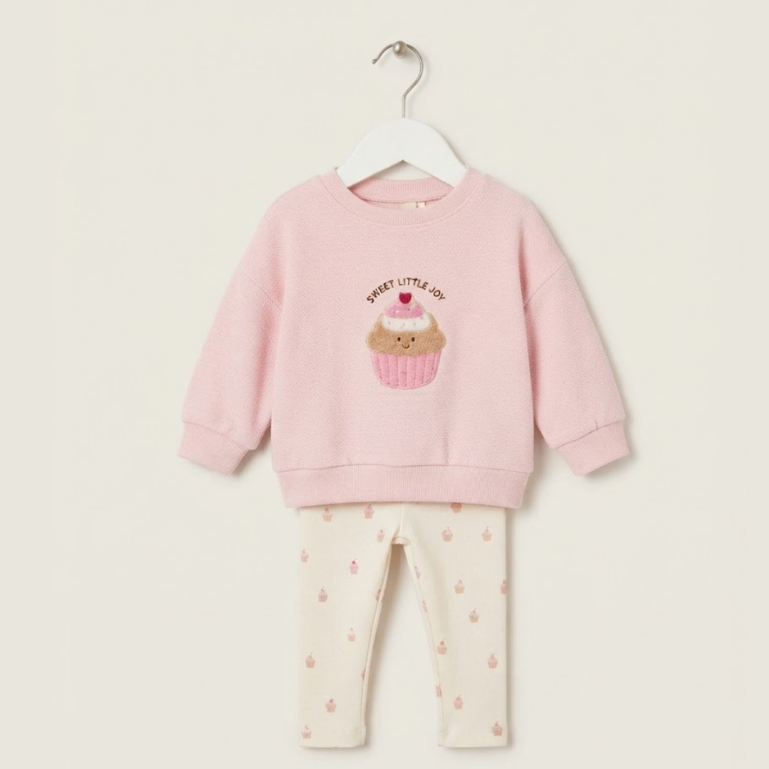 A toddler girl's two-piece pajama set hanging on a white hanger against a light beige background. The set includes a light pink long-sleeve sweatshirt with a cute cupcake graphic and the text 'SWEET LITTLE DOG' on the front, paired with cream-colored leggings patterned with small pink hearts.