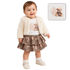 A baby girl wearing a cream faux-fur cardigan, a white t-shirt with a teddy bear graphic, and a brown leopard-print tiered skirt, standing against a white background. An inset image in the top right shows a matching teddy bear plush toy.