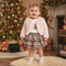 A toddler girl stands in a decorated Christmas room wearing a light pink fuzzy cardigan over a white graphic t-shirt and a brown leopard-print tiered skirt with pink bows. She is smiling and posing in front of a lit Christmas tree and fireplace mantel.