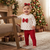 A toddler stands barefoot in a living room near a Christmas tree, wearing a white long-sleeve top with small red motifs and a large red bow at the chest, paired with red leggings and a red bow headband.
