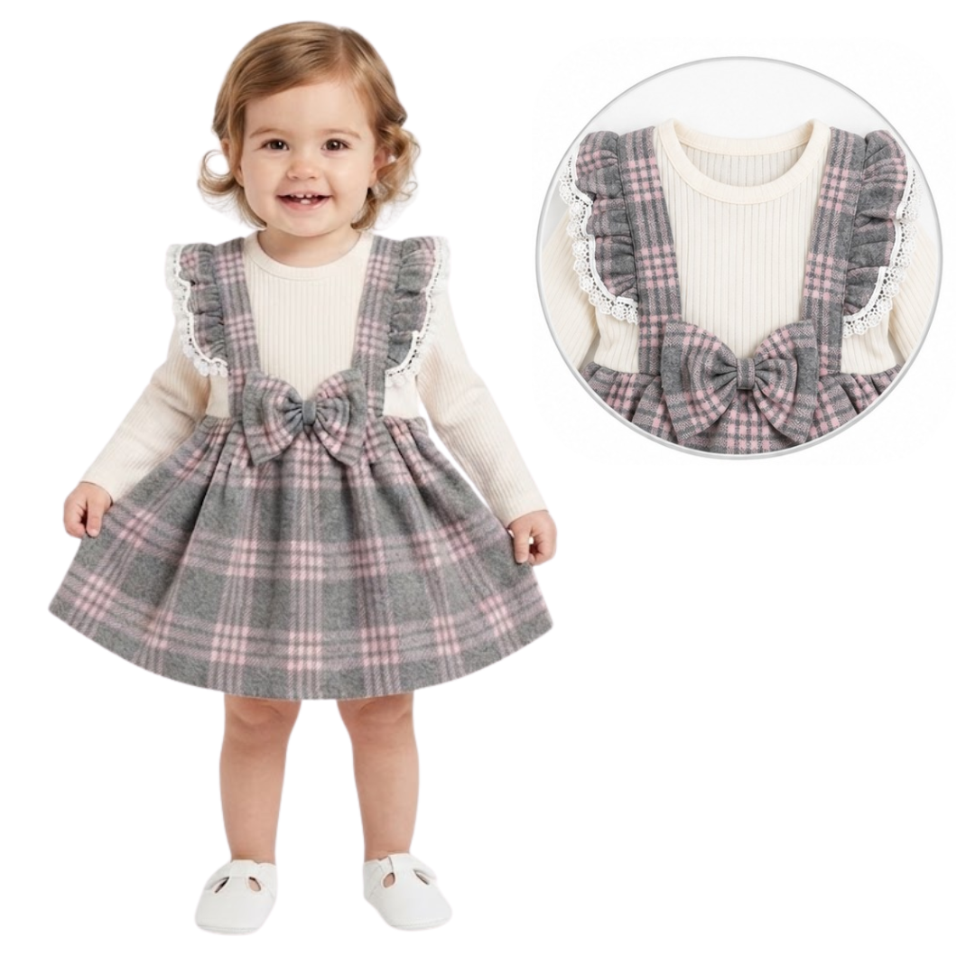 A toddler stands against a white background wearing a gray plaid pinafore-style dress over a cream ribbed long-sleeve top. The dress has ruffled shoulder straps, a large bow at the waist, and a flared skirt.