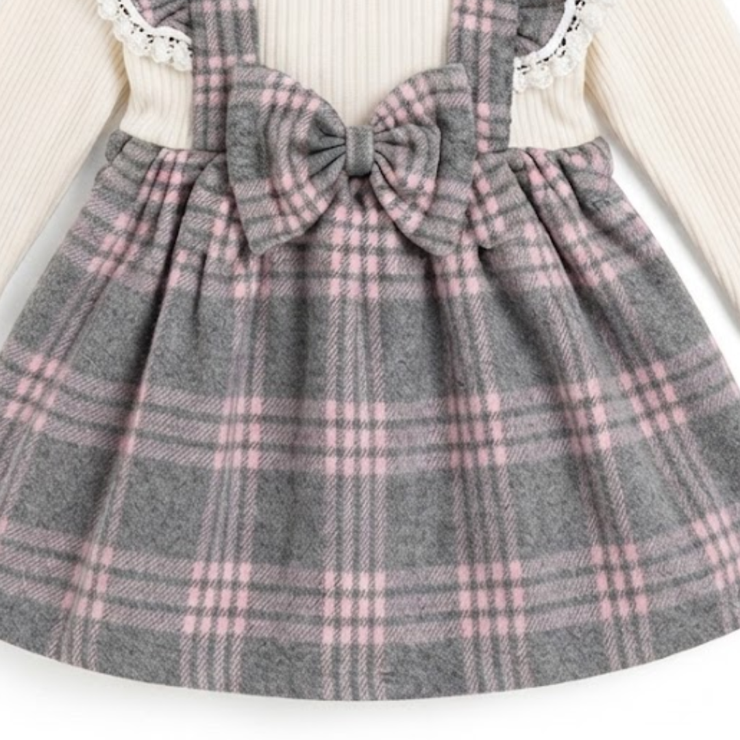 A close-up of a child's dress featuring a gray and pink plaid skirt with a large matching bow at the center, layered over a cream ribbed long-sleeve top with white lace trim.