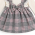 A close-up of a child's dress featuring a gray and pink plaid skirt with a large matching bow at the center, layered over a cream ribbed long-sleeve top with white lace trim.