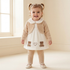 A toddler girl with blonde pigtails stands smiling in a bright cream room, wearing a cream and beige outfit consisting of a long-sleeve knit cardigan, a white dress with teddy bear and heart embroidery, matching cream leggings, and cream shoes.