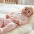 A smiling baby lying on a white faux fur blanket, wearing a light pink footed sleeper with a smocked chest design.