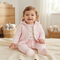 A baby girl wearing a light pink quilted jacket and matching pink footed pants sits smiling on a cream knit blanket in a softly lit nursery.