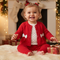 A baby wearing a festive red Christmas outfit with a matching beanie, lace trim, and white bow, sitting on a cream knit chair in front of a decorated Christmas tree.